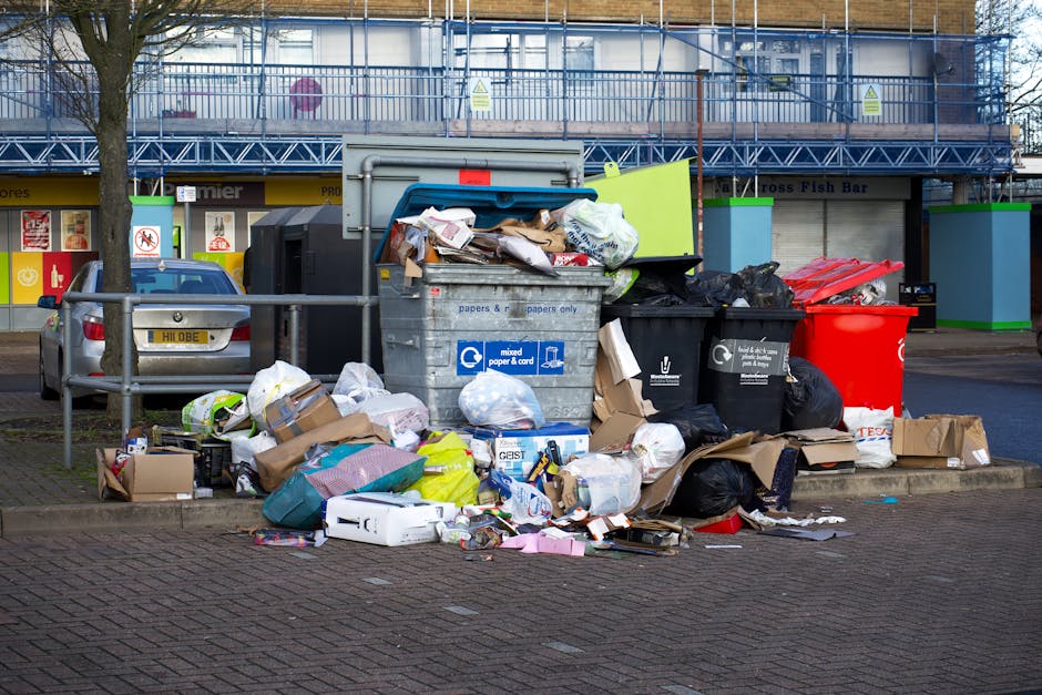 A large collection of mixed household waste and rubbish overflowing from a central wheeled bin on a paved sidewalk in an urban area. The bin, made of metal with a blue recycling label, is open and filled with crumpled paper, cardboard boxes, plastic bags, and other domestic waste materials. Surrounding the bin are additional black and red rubbish bags, flattened cardboard boxes, and loose paper debris scattered on the ground. Behind the pile, there are parked vehicles, including a grey car with a yellow registration plate. In the background, a commercial storefront with brightly colored signage, indicating retail shops or food outlets, is visible along with a building under scaffolding, suggesting ongoing construction or maintenance. The scene is illuminated by natural daylight, with a moderate level of urban activity typical of an area where private waste disposal and rubbish removal services like those offered by Rubbish Removal Hounslow are commonly used for managing waste outside of council collection services.