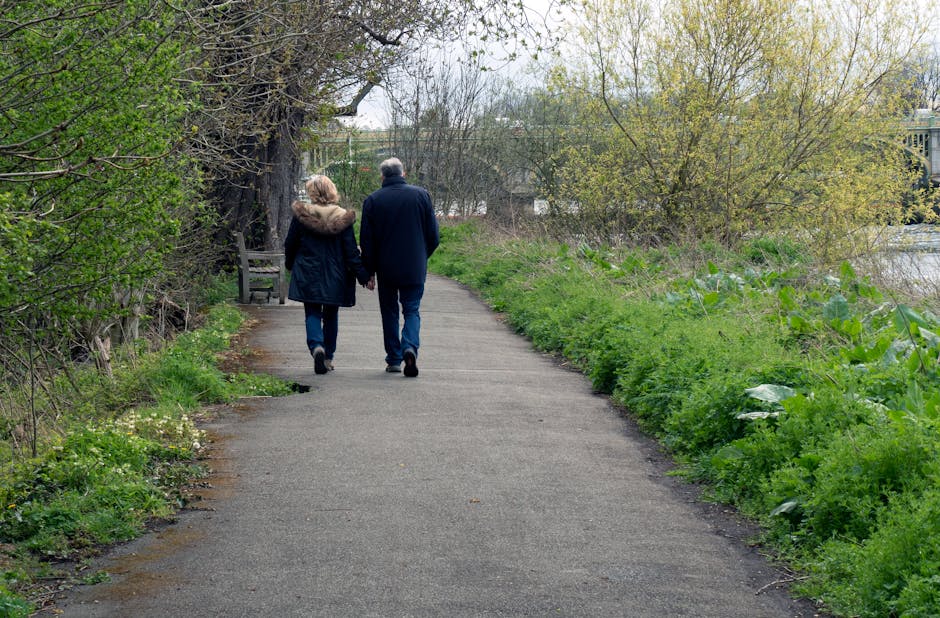 A photograph depicts a paved footpath in a natural setting during daytime, where an elderly couple is walking away from the camera, holding hands. The woman is dressed in a dark coat with a fur-lined hood, while the man is wearing a navy blue jacket and jeans. The path is bordered by lush green grass, shrubs, and small wildflowers on the left, and a dense cluster of trees and bushes with new spring foliage on the right. In the background, more trees and a glimpse of water or a river can be seen, with some buildings partially visible beyond the foliage. The scene appears calm and quiet with soft natural lighting, emphasizing the peaceful outdoor environment suitable for private outdoor activities and independent leisure walks. Rubbish Removal Hounslow might facilitate waste collection in such natural areas, ensuring the preservation of these environments for community use.