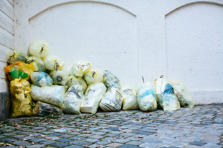 A collection of tightly packed plastic rubbish bags, predominantly in shades of yellow and white, are arranged along the base of a white, textured exterior wall. The bags are made of thin, semi-transparent plastic, allowing glimpses of discarded packaging and waste inside. Some bags are knotted at the top with black ties, while others are loosely sealed or folded over. The ground beneath the bags consists of cobblestone paving with small, irregularly shaped stones in gray and brown tones, with a few scattered leaves and debris nearby. The backdrop features a smooth, white wall with subtle arched indentations, suggesting an external boundary of a property or building. The setting appears to be an outdoor urban alley or pathway, where waste has been temporarily stored, likely awaiting collection by private rubbish removal services such as Rubbish Removal Hounslow, especially in the context of alternative on-site waste disposal or booking challenges for rubbish collection in the area.