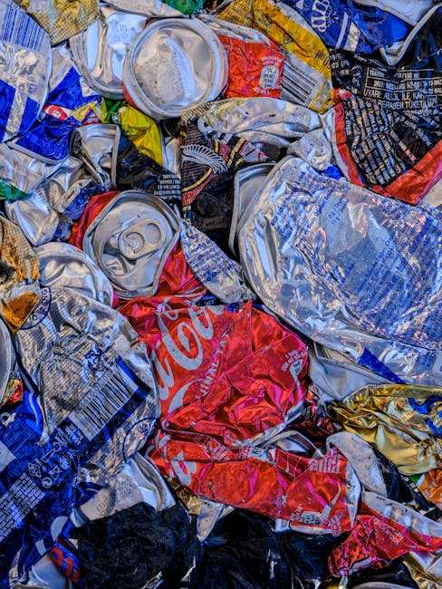 A close-up view of a pile of crushed and crumpled aluminium cans, predominantly in silver, with visible branding and logos, including a partially visible red Coca-Cola can. The cans display various textures, from shiny metallic surfaces to matte and torn areas, with some featuring pull-tabs and ridges. The background shows more flattened cans tightly packed together, creating a cluttered, dense arrangement. Natural light highlights the reflective qualities of the aluminium, emphasizing the metallic sheen and the weathered appearance of some cans. The scene suggests a typical waste collection or recycling process, possibly handled by a waste removal service such as Rubbish Removal Hounslow, which specialises in rubbish and waste management. The cans are contained within a large collection, suitable for either disposal or recycling, and the close proximity of the objects indicates an on-site collection or pre-sorting for waste removal. This detailed description offers insights into metal waste items, aligning with the context of alternative waste handling methods away from local authority bulk rubbish collection.