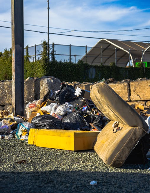 A collection of discarded rubbish piled on the ground in an outdoor area, including black and white plastic rubbish bags, a yellow plastic container, and an old, worn car tire tilted on its side. The rubbish is set against a low stone wall composed of roughly cut, weathered stones with visible mortar. In the background, there is a dark green hedge, metal fencing, and a partially visible large, curved bus shelter structure with a beige roof, situated on a paved surface. Overhead, power lines run across a partly cloudy sky with patches of blue. The scene suggests an informal waste disposal site near a public space or roadway, consistent with the context of independent rubbish removal services in Hounslow. Rubbish Removal Hounslow occasionally handles such bulk accumulations as part of their rubbish collection methods, which differ from official council waste disposal channels.