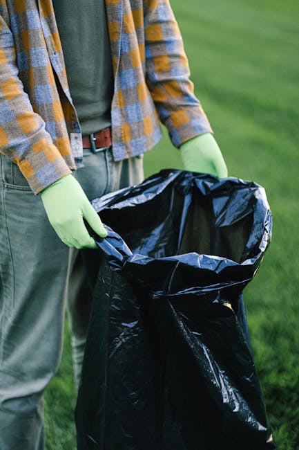 A person wearing a checkered yellow and gray shirt, gray trousers with a brown belt, and light green gloves is holding open a large black plastic rubbish bag, preparing for waste disposal. The scene is set outdoors on a grassy area, with vibrant green grass visible in the background. The individual’s hands are positioned at the top of the bag, which appears to be partially collapsed and ready for collection of waste. This activity aligns with rubbish removal or waste management services, highlighting on-site waste collection as an alternative to scheduled council rubbish pickups, indicative of private disposal or independent waste handling. The focus of the image emphasizes the manual process of rubbish collection, with the surrounding environment providing a natural, outdoor setting consistent with garden or outdoor rubbish clearance tasks, often undertaken by professional rubbish removal services such as Rubbish Removal Hounslow.