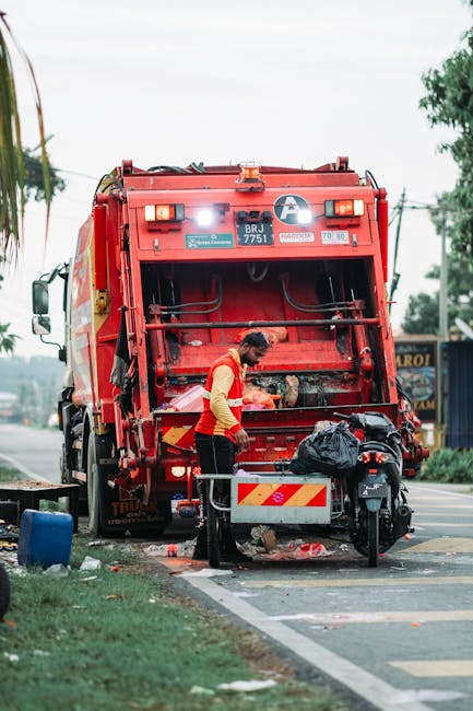 A worker wearing a red and yellow uniform is engaged in rubbish collection, standing beside the open rear compartment of a large red waste collection truck on a roadside. The truck, branded with identification labels, has its rear hatch lifted, revealing the storage area inside. The worker appears to be loading or securing waste, with a black trash bag placed on a nearby cart or platform attached to the vehicle. The scene takes place on an urban street, with a sidewalk, some greenery, and distant trees visible in the background. The environment is well-lit with natural daylight, and the surrounding area includes utility poles and signs, indicating an active roadside environment. The image clearly depicts private waste management activity, likely an alternative to municipal rubbish removal services, illustrating on-site or independent rubbish collection employed by companies such as Rubbish Removal Hounslow.