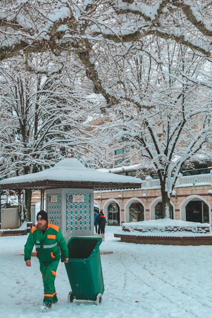 A waste collection worker dressed in a green uniform with orange reflective stripes is walking on a snow-covered pavement, pushing a large green wheeled rubbish bin. Behind him, there is a small, tiled shelter with a curved roof, also covered in snow. Several leafless trees with snow-laden branches tower in the background, alongside a brick wall with white arches and decorative tiles. A person wearing a dark coat and red hat is seen walking in the distance. The whole scene is set in a wintery outdoor environment with snow blanketing the ground, trees, and structures, suggesting an outdoor public space such as a park or town square. The setting reflects a typical rubbish collection or waste management activity taking place during winter, emphasizing independent collection services provided by companies like Rubbish Removal Hounslow.
