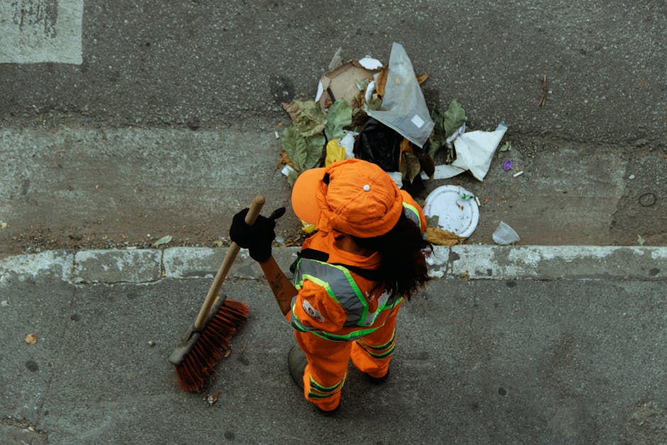 A waste collection worker dressed in a green uniform with orange reflective stripes is walking on a snow-covered pavement, pushing a large green wheeled rubbish bin. Behind him, there is a small, tiled shelter with a curved roof, also covered in snow. Several leafless trees with snow-laden branches tower in the background, alongside a brick wall with white arches and decorative tiles. A person wearing a dark coat and red hat is seen walking in the distance. The whole scene is set in a wintery outdoor environment with snow blanketing the ground, trees, and structures, suggesting an outdoor public space such as a park or town square. The setting reflects a typical rubbish collection or waste management activity taking place during winter, emphasizing independent collection services provided by companies like Rubbish Removal Hounslow.