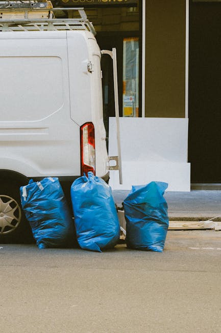 A white commercial van is parked on a paved street near the entrance of a building with large glass doors. The van's rear door is closed, and the vehicle features a red tail light and a side mirror. In front of the van, on the pavement, are three blue rubbish bags made of plastic, filled and tied at the top, with slight creases and folds visible on their surfaces. The bags appear to contain mixed waste material, and are positioned side by side, leaning slightly into each other. The building's exterior includes a white decorative wall element and a small step leading to the entrance, suggesting a private property or commercial premises. The scene is illuminated by natural daylight, providing clear visibility of the textures—the smooth, glossy finish of the van's paint, the crinkled plastic bags, and the clean, even pavement. This setup reflects an alternative rubbish collection method, possibly involving private waste handling or on-site clearance, with Rubbish Removal Hounslow offering similar services for immediate rubbish disposal and collection needs.