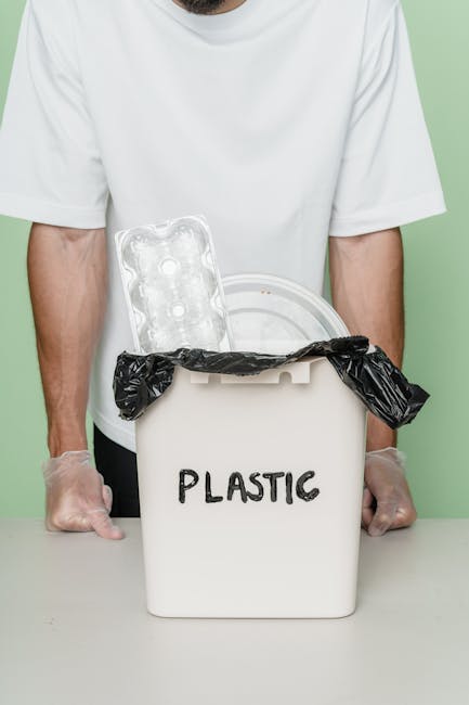 A person wearing a white t-shirt and transparent disposable gloves is standing behind a white plastic bin labeled 'PLASTIC' in black lettering. The bin has a black plastic bag lining it, with the excess bag hanging over the edges. Resting on top of the bin is an empty, clear plastic blister pack, positioned next to the open lid. The background features a light green wall, and the scene appears to be indoors, possibly in a collection or waste management setting. The image visually emphasizes the segregation and collection of plastic waste, aligning with independent rubbish collection practices and on-site disposal methods supported by services like Rubbish Removal Hounslow.