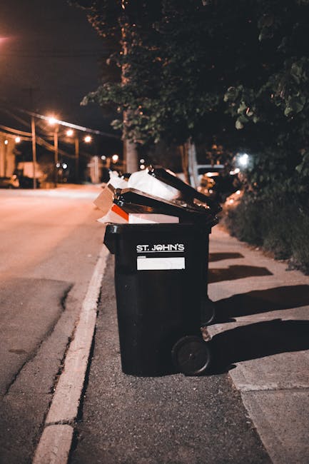 A person wearing a white t-shirt and transparent disposable gloves is standing behind a white plastic bin labeled 'PLASTIC' in black lettering. The bin has a black plastic bag lining it, with the excess bag hanging over the edges. Resting on top of the bin is an empty, clear plastic blister pack, positioned next to the open lid. The background features a light green wall, and the scene appears to be indoors, possibly in a collection or waste management setting. The image visually emphasizes the segregation and collection of plastic waste, aligning with independent rubbish collection practices and on-site disposal methods supported by services like Rubbish Removal Hounslow.