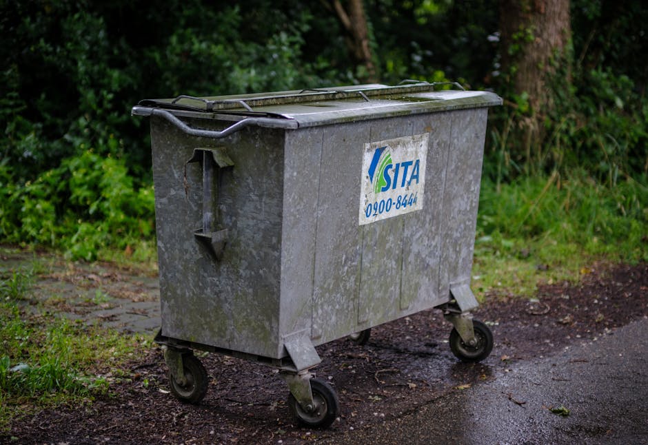 A large, industrial-style wheeled rubbish bin made of weathered, gray metal with visible dirt and rust spots, positioned on a pavement near a grassy area with dense green foliage and trees in the background. The bin features a sealed, flat lid with a raised handle on the side for lifting, and a manufacturer's label with the logo and phone number of a waste collection service. The surrounding environment includes a concrete or asphalt surface, with some patches of dirt and moss, indicative of outdoor use in a residential or semi-urban setting. The scene suggests an outdoor waste disposal point where private waste collection services, such as those provided by Rubbish Removal Hounslow, may carry out rubbish removal or on-site clearance, emphasizing the importance of proper waste management and adherence to local waste laws. Natural lighting highlights the textured surface and the mixture of vegetation and pavement, creating a neutral, observation-focused image.