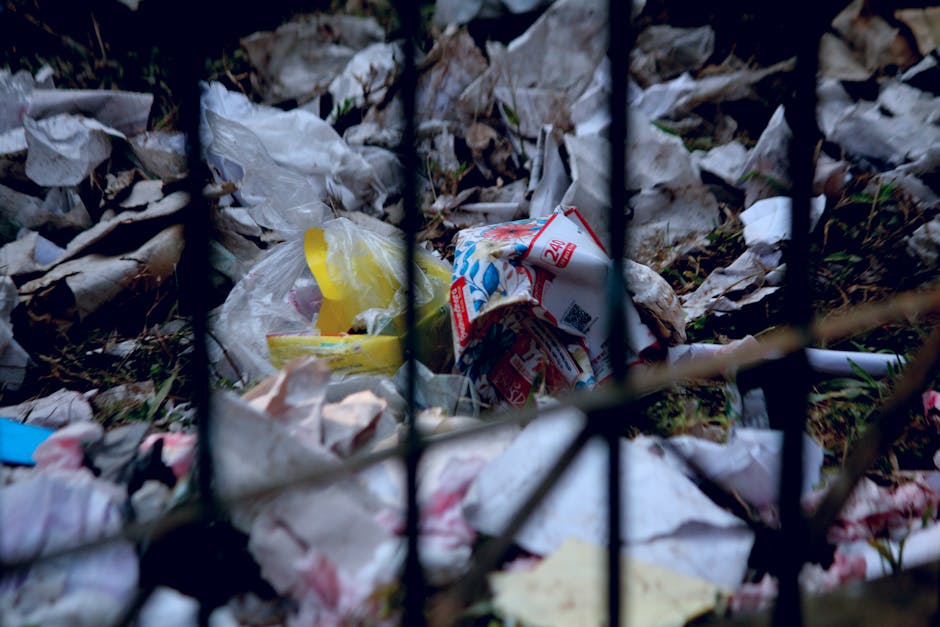 A patch of ground and grassy slope scattered with various types of discarded packaging materials, including flattened cardboard boxes with printed logos and open containers that appear to have previously held beverages. Some boxes are crushed, torn, or partially collapsed, with the cardboard displaying a mix of brown and greenish tones, and the printed logos featuring white star symbols and text. The debris is irregularly spread across the area, with a few larger pieces near the center and smaller fragments dispersed throughout. A thin metal or plastic pipe runs diagonally through the scene, partially covered by the rubbish. In the background, there is a wooden fence or wall, suggesting this scene is outside, possibly along a roadside or in a neglected outdoor space. The environment appears unkempt, with sparse grass and dried vegetation on the slope, and the lighting is natural, indicating daytime with no visible shadows or highlights. This scene illustrates a typical example of illicit waste dumping that local rubbish removal services might assist with in Hounslow.